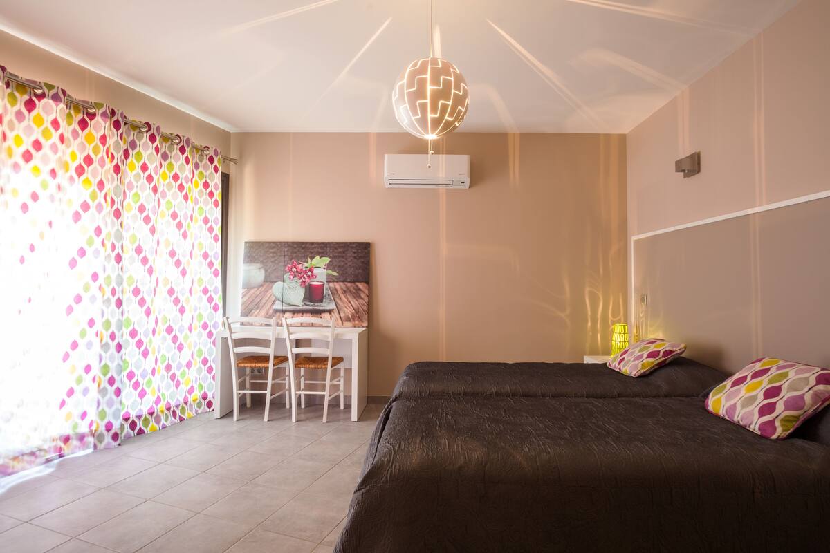 A bedroom with a bed, a desk with a chair, a window with curtains, and a wall-mounted air conditioner.