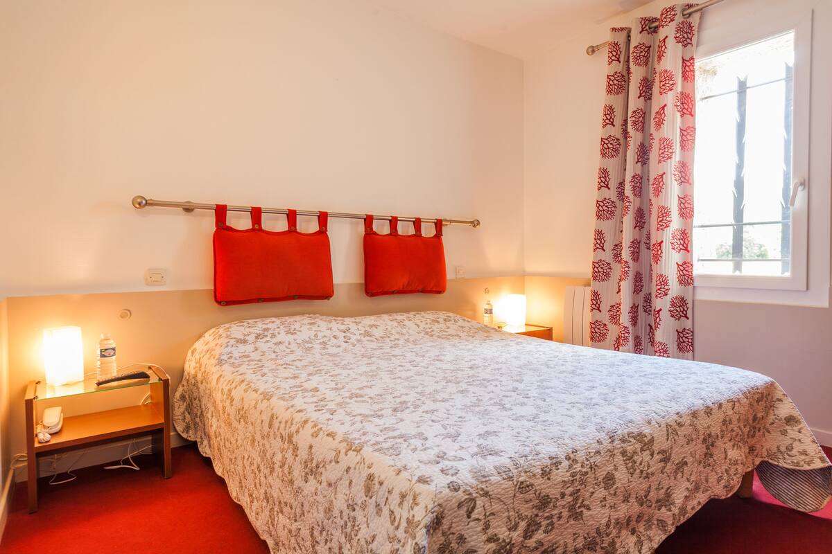 A bedroom with a floral bedspread, two red pillows, a bedside table with a lamp, and a window with patterned curtains.