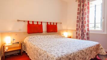 Un dormitorio con colcha estampada, dos almohadas rojas, una mesita de noche con lámpara y una ventana con cortinas decoradas.