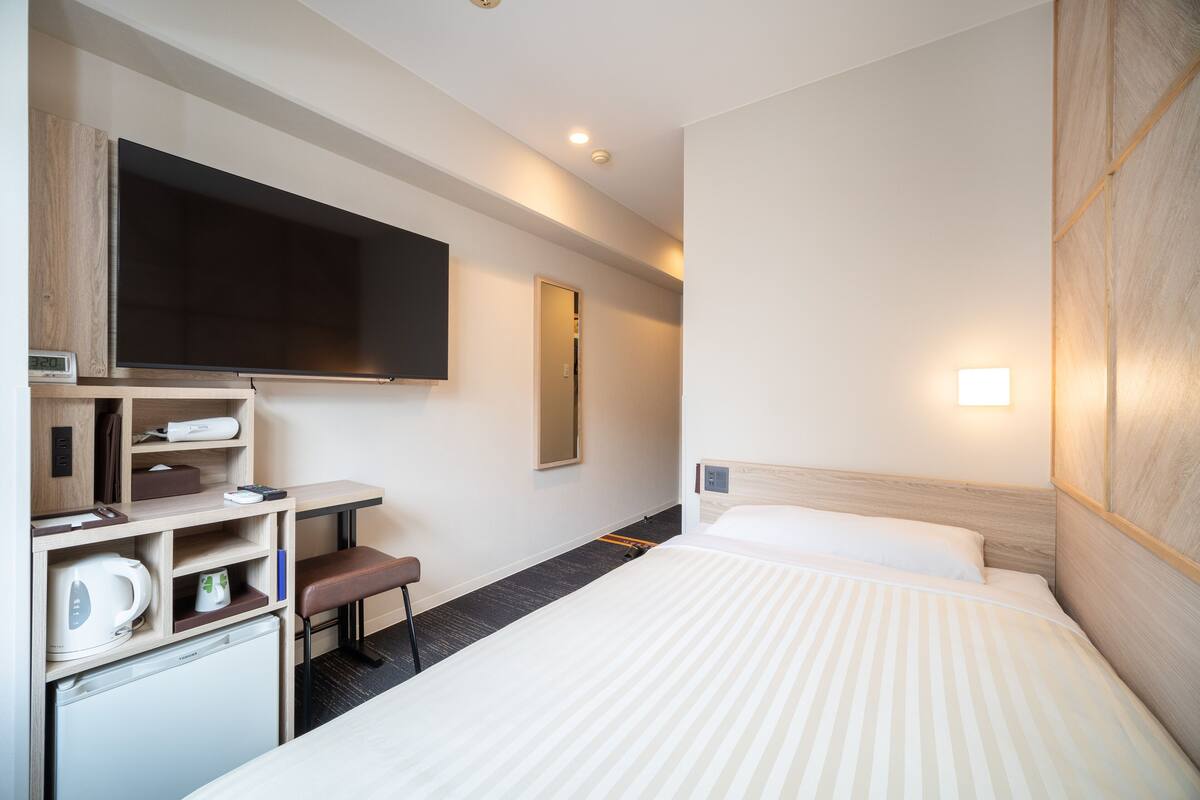 A hotel room with a bed, a TV, a desk, a chair, and a small kitchenette.