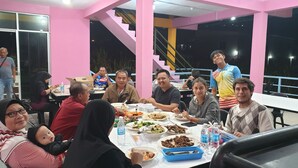 A group of people dining at a table in a brightly colored room with pink walls and a staircase in the background. - Kupi-Kupi & Stay (Ranau)