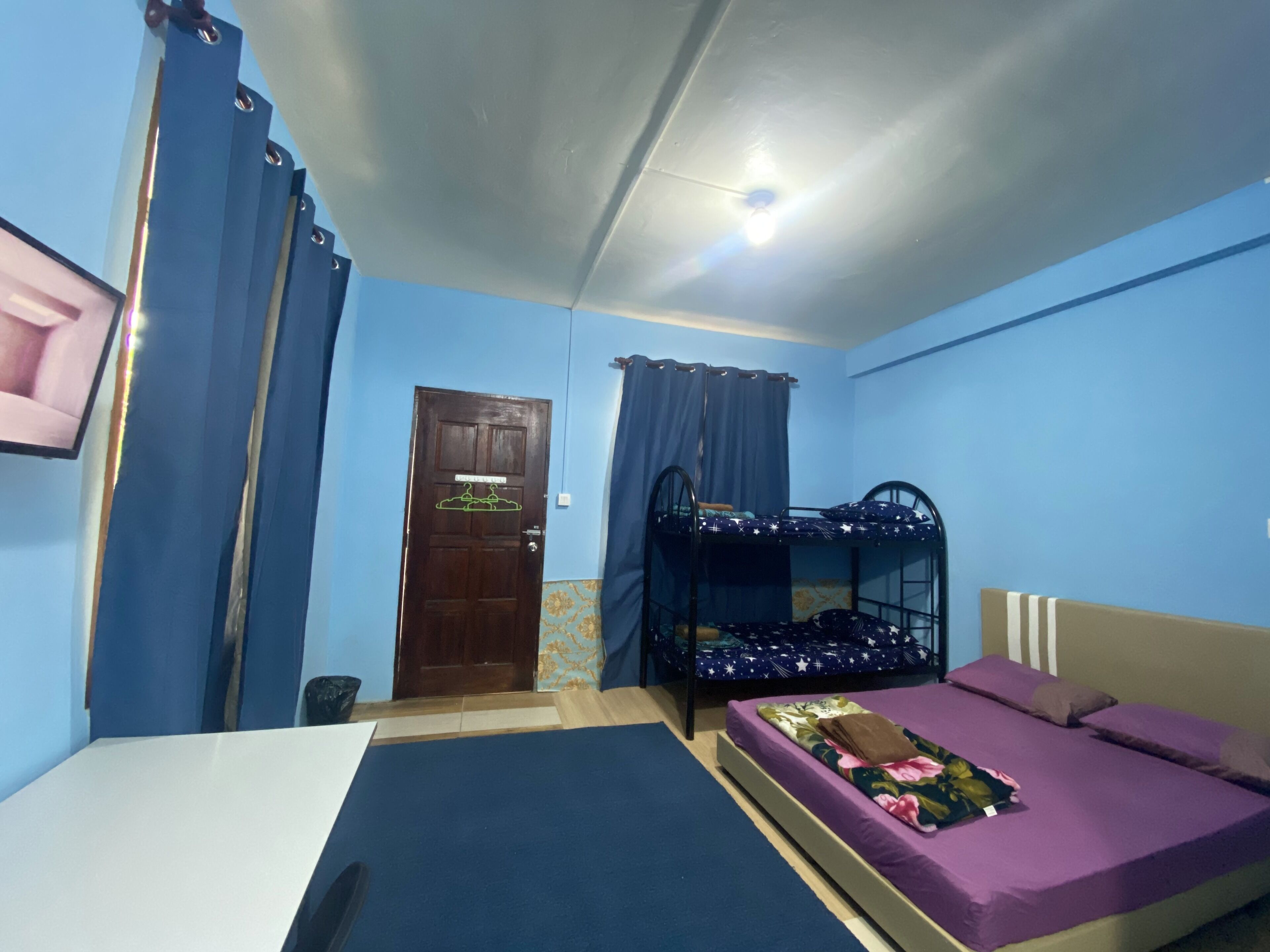 Een kamer met een stapelbed, een eenpersoonsbed, een deur en blauwe gordijnen.