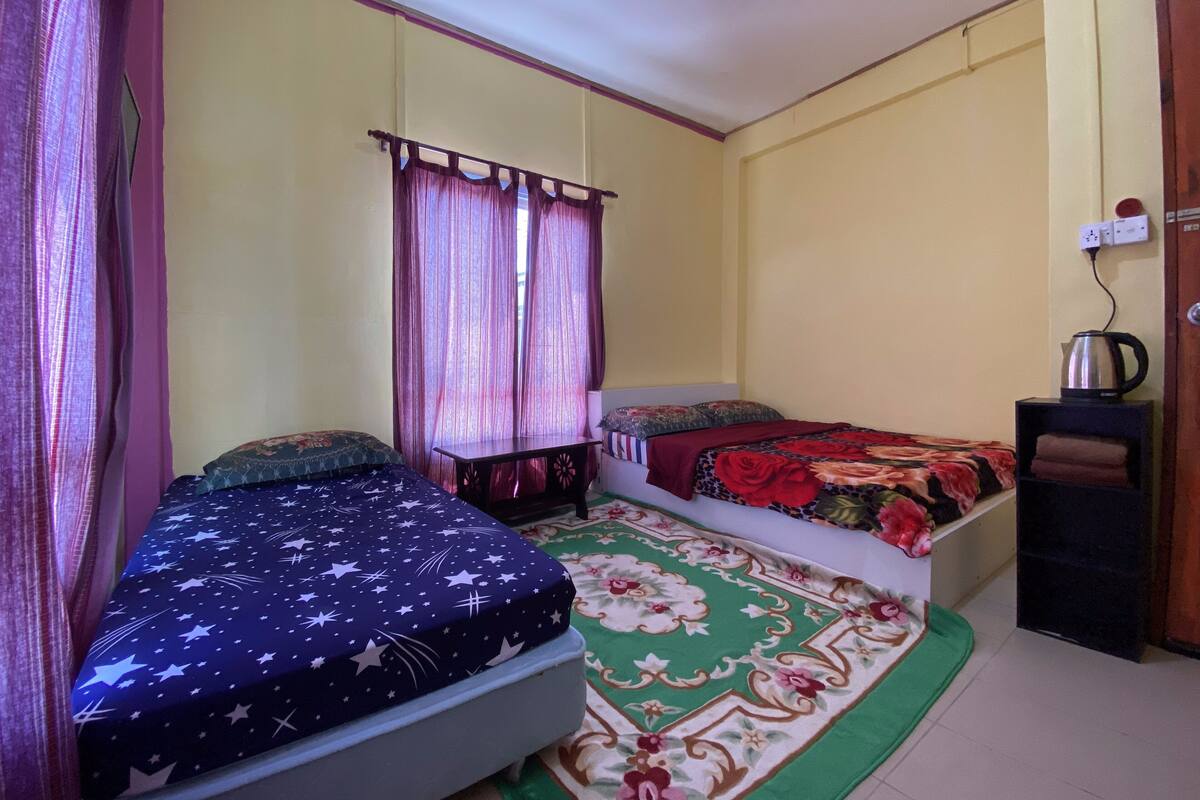 A bedroom with two single beds, a small table, a chair, a window with curtains, and a kettle on a shelf.