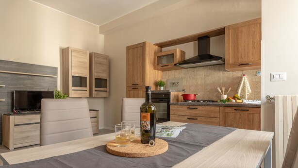 A modern kitchen with wooden cabinets, a dining table with a cork coaster, a bottle of wine, and a TV on a stand. - Manzoni Apartment (Roseto degli Abruzzi)