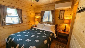 Ein gemütliches Holzblockhaus-Zimmer mit einem Bett, zwei mit Vorhängen versehenen Fenstern, einem Deckenventilator, einem Nachttisch mit Lampe und einer wandmontierten Klimaanlage.