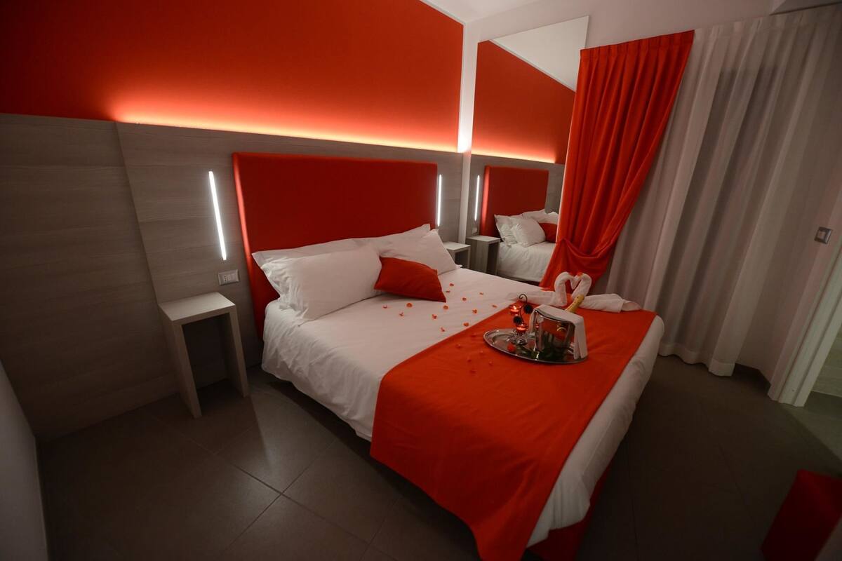 Una habitación de hotel con una cama grande, detalles en rojo, una mesita de noche y un espejo.