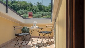 A small balcony with a table and two chairs, a potted plant, and a view of greenery.