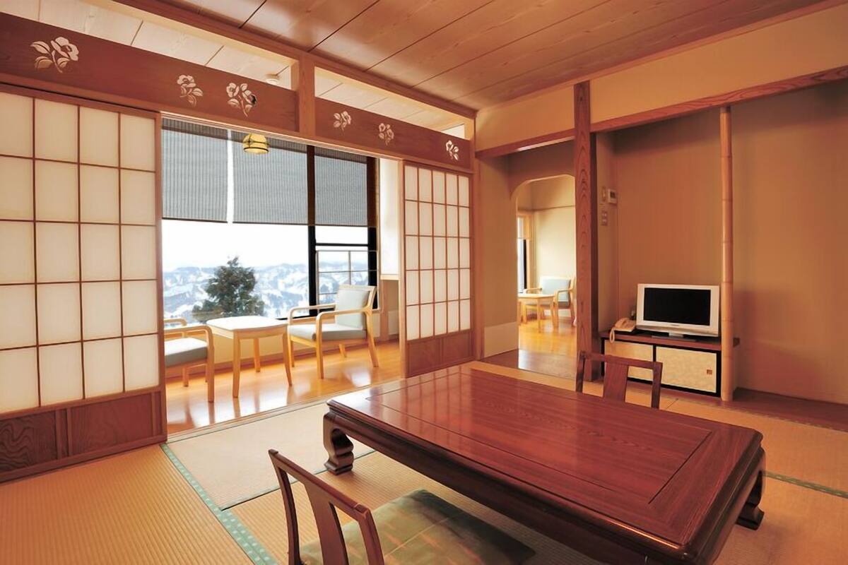 Een traditionele Japanse kamer met tatami vloeren, een houten tafel, een televisie en een schuifdeur naar een balkon met uitzicht.