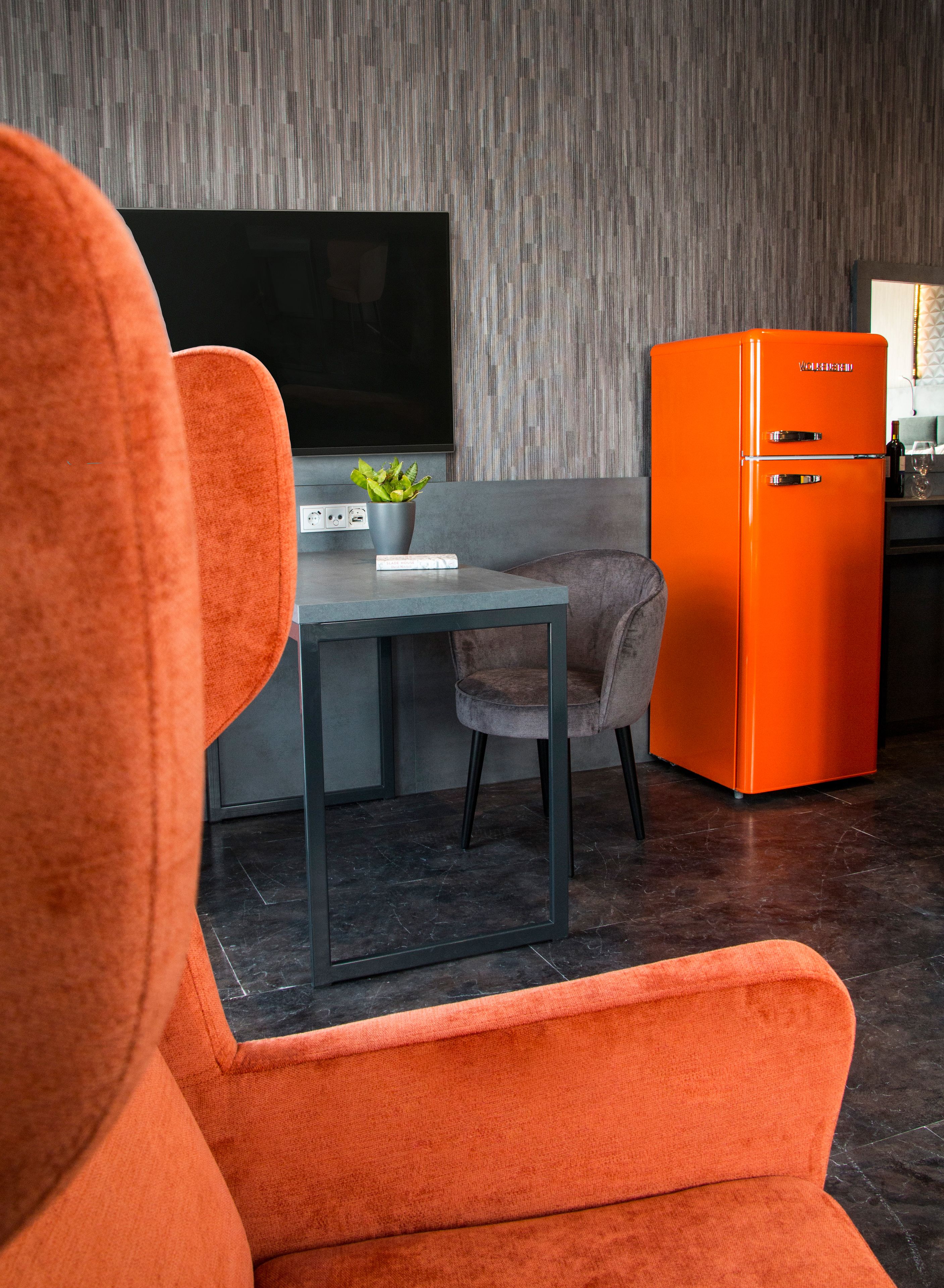 Una sala moderna con un sofá gris, una mesita con una planta y un refrigerador naranja brillante.