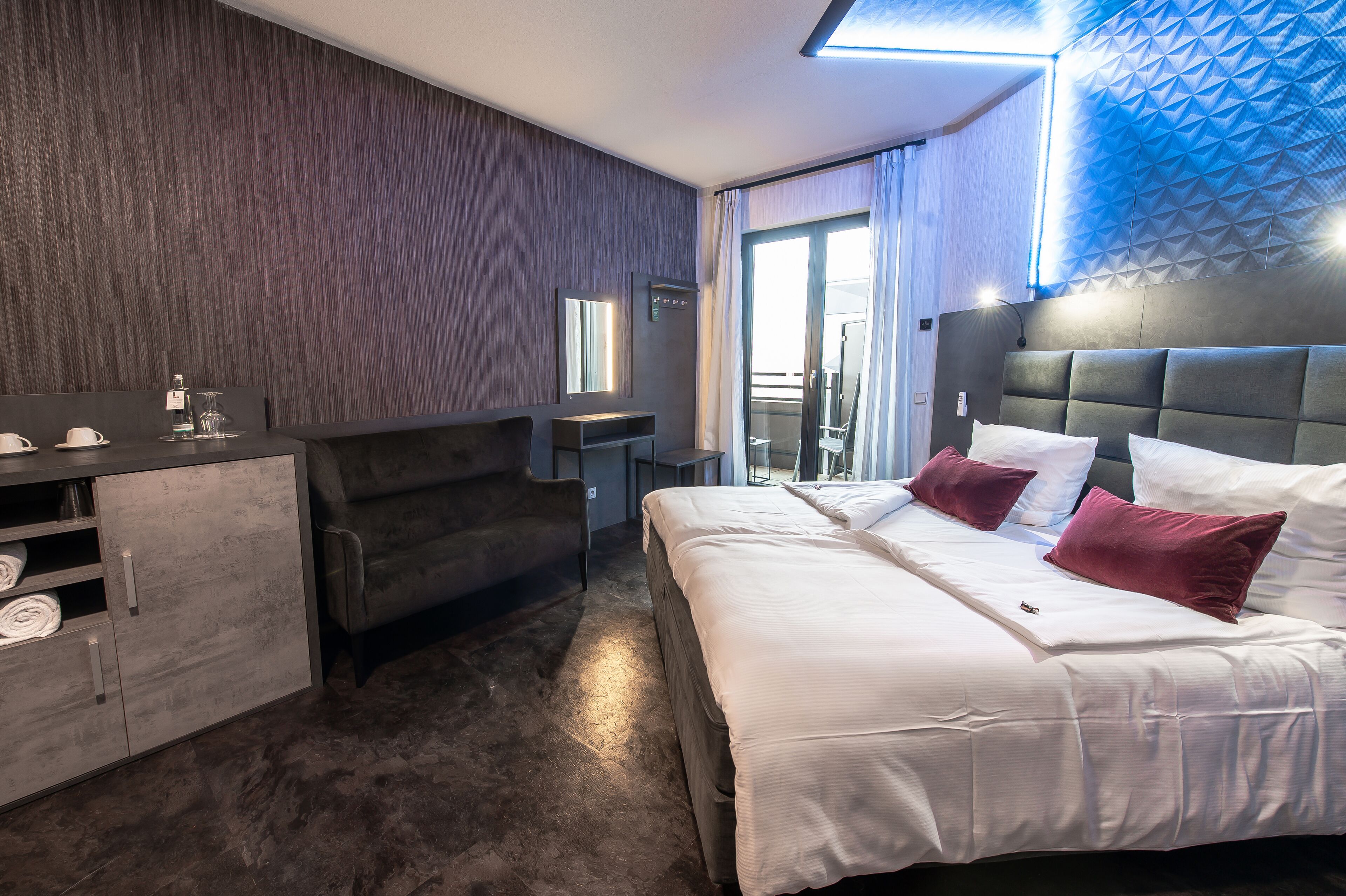 A modern hotel room with a large bed, a sofa, a desk, and a wardrobe.
