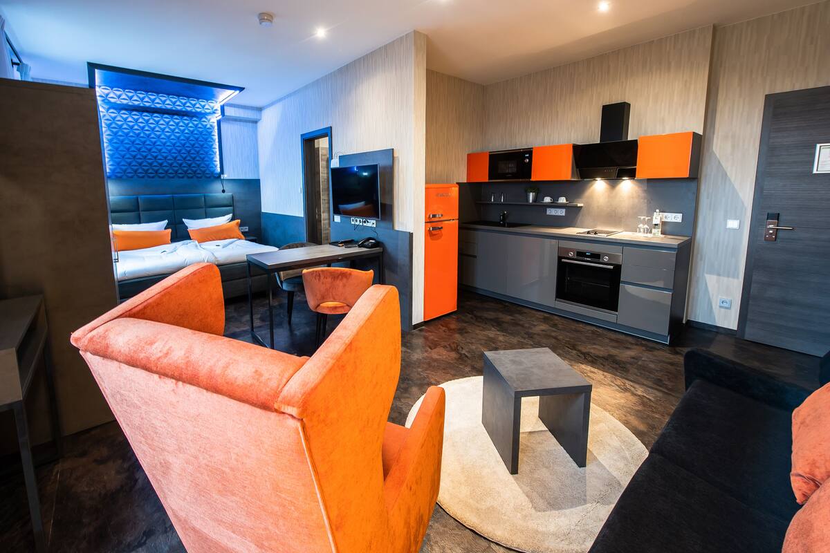 A modern hotel room with a bed, orange armchair, small table, and a kitchenette with a microwave and oven.