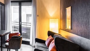 Una habitación de hotel moderna con una pared de madera oscura, un sofá con cojines naranja, una mesa de comedor con sillas y un amplio ventanal con cortinas.