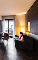 Quarto de hotel moderno com uma parede de madeira escura, um sofá com almofadas laranja, uma mesa de jantar com cadeiras e uma janela grande com cortinas.