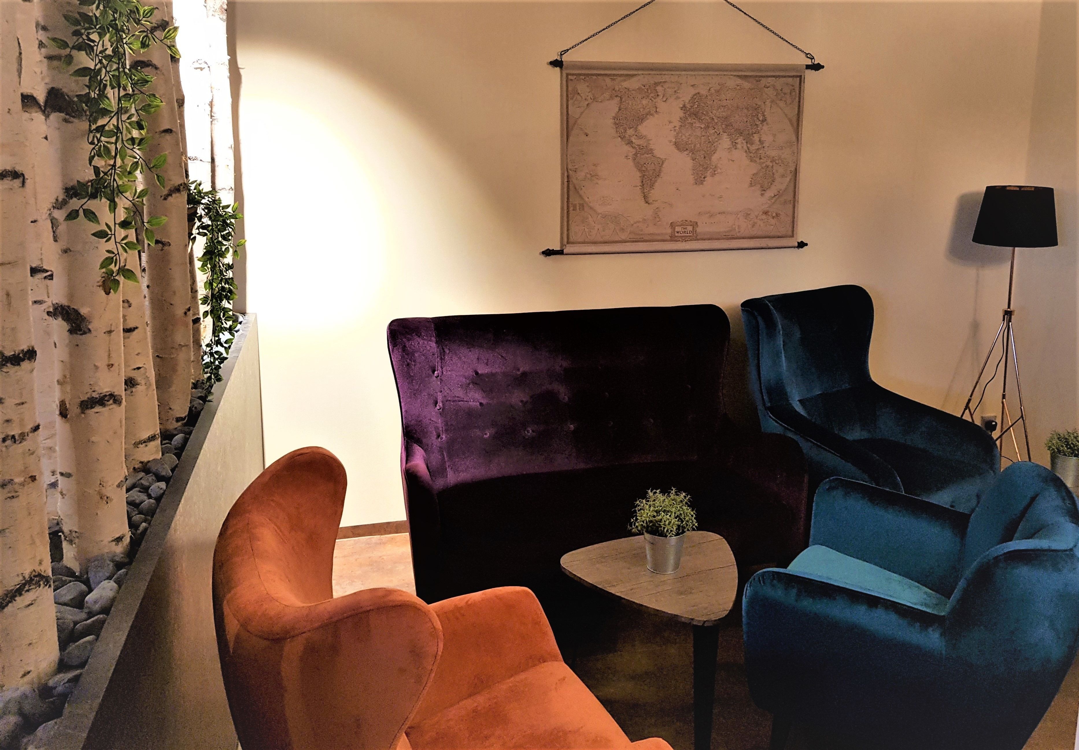 A room with a large purple sofa, a smaller orange armchair, and a blue armchair. There is a small table with a plant in the center. A world map hangs on the wall above the sofa.