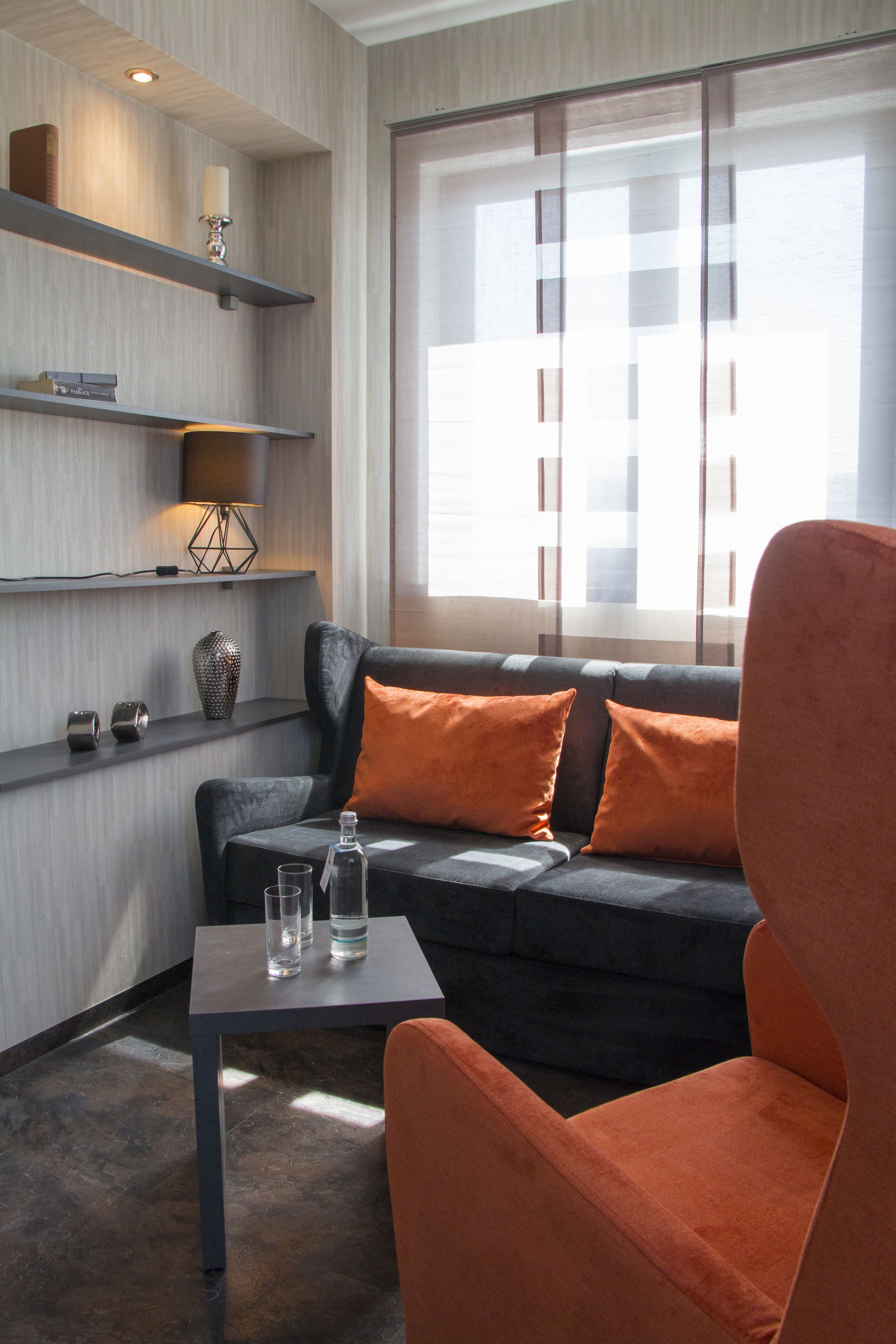 Un salon moderne avec un canapé, des coussins orange, une table avec des verres et une bouteille, et des étagères ornées d’objets décoratifs.