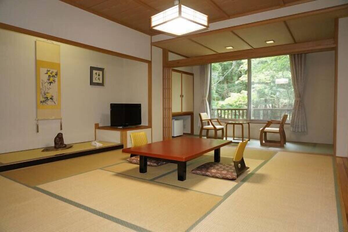 Ein traditioneller japanischer Raum mit Tatami-Böden, einem niedrigen Tisch, Stühlen, einem Fernseher und Blick auf Grünflächen.