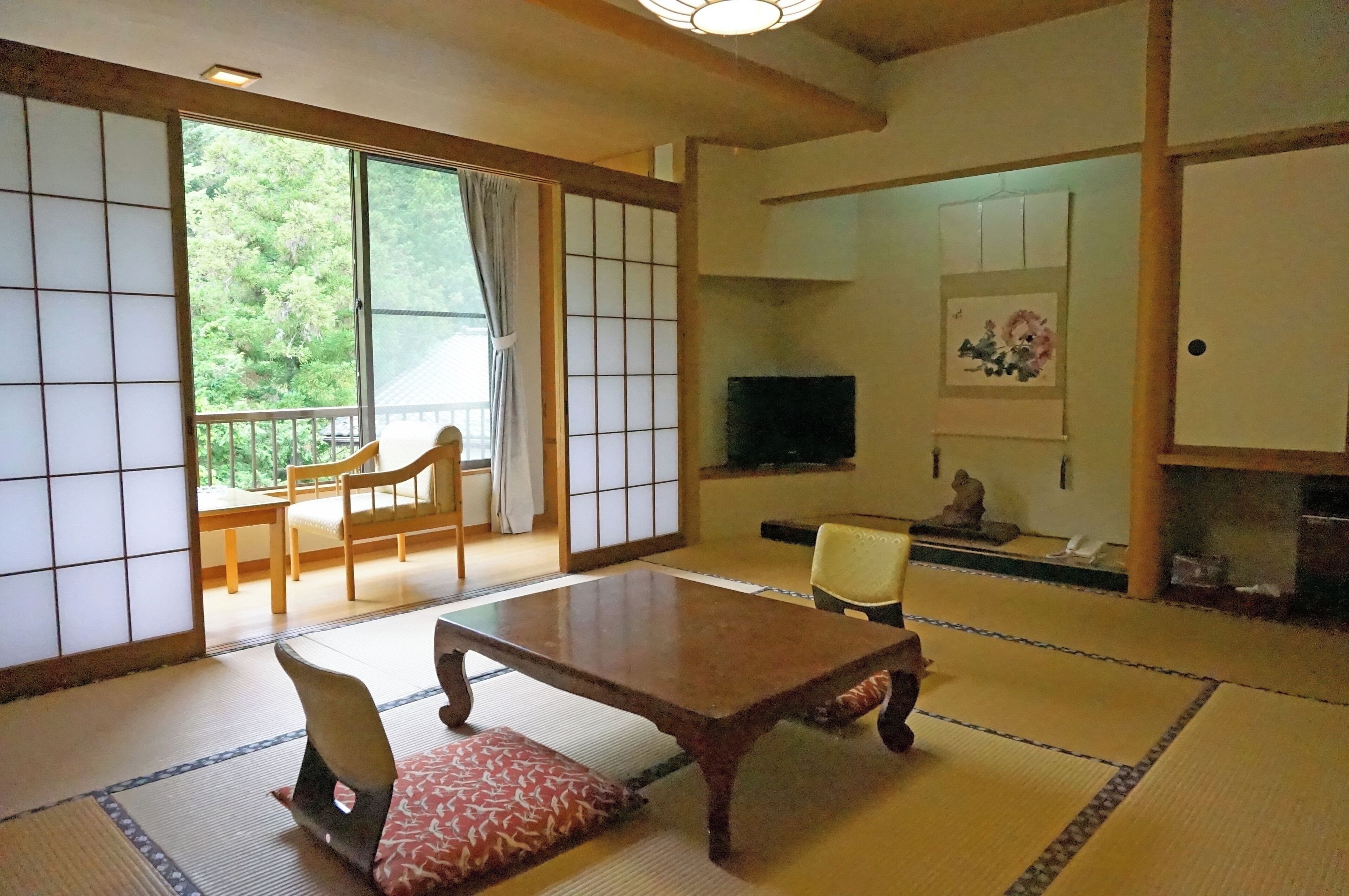 Una habitación japonesa tradicional con suelo de tatami, una mesa baja, sillas, un televisor y una puerta corrediza que da a un balcón.