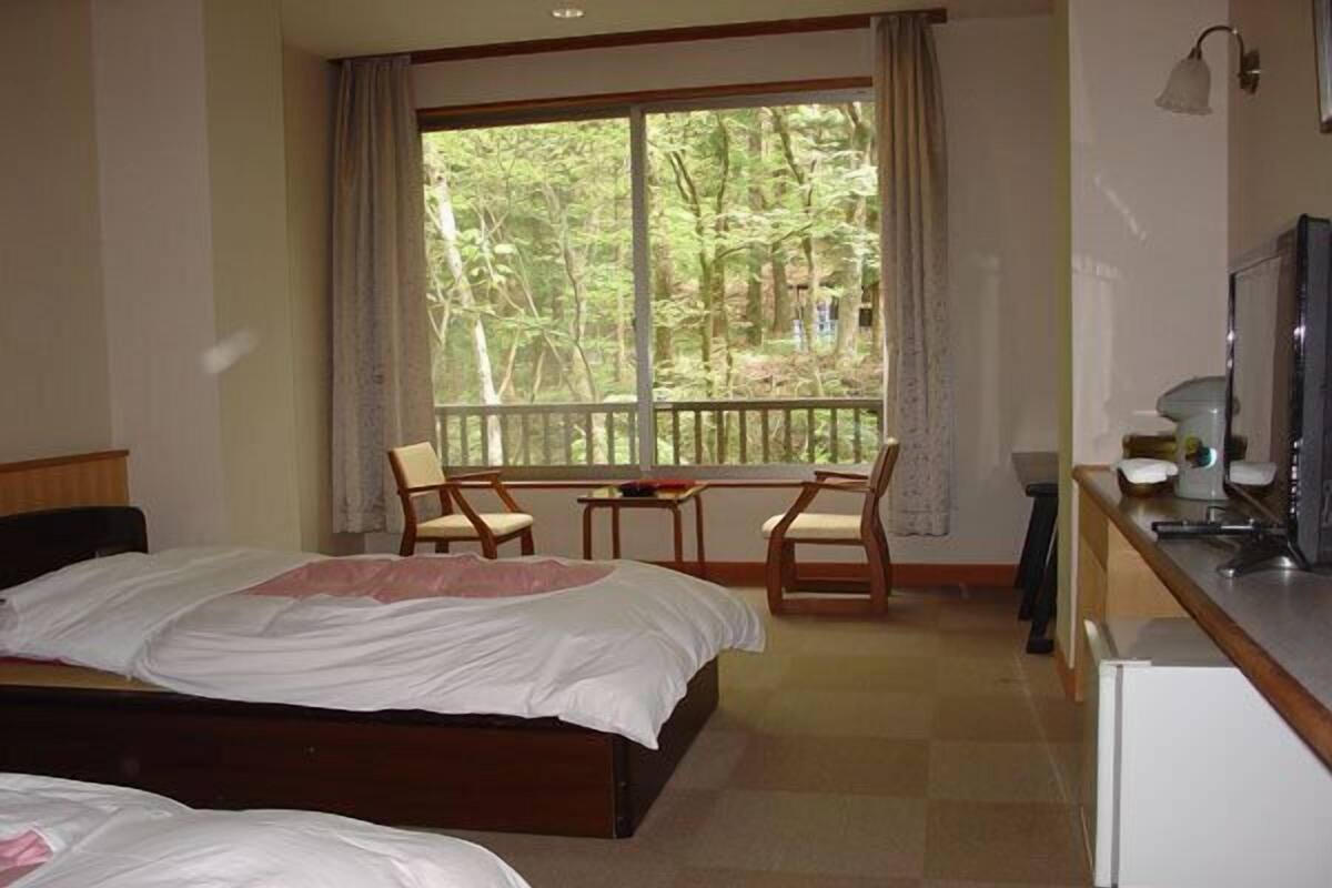 Ein Schlafzimmer mit zwei Betten, einem kleinen Tisch, zwei Stühlen, einem Schreibtisch mit Computer und einem Fenster mit Blick auf Bäume.