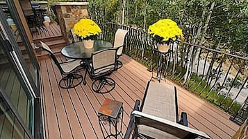 A wooden deck with a glass-top table and chairs, surrounded by a metal railing and greenery.