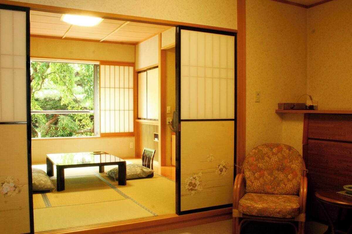 Een traditionele Japanse kamer met tatami vloerbedekking, een verschuifbaar shoji-scherm, een lage tafel met een kussen en een stoel met een bloemenprint.