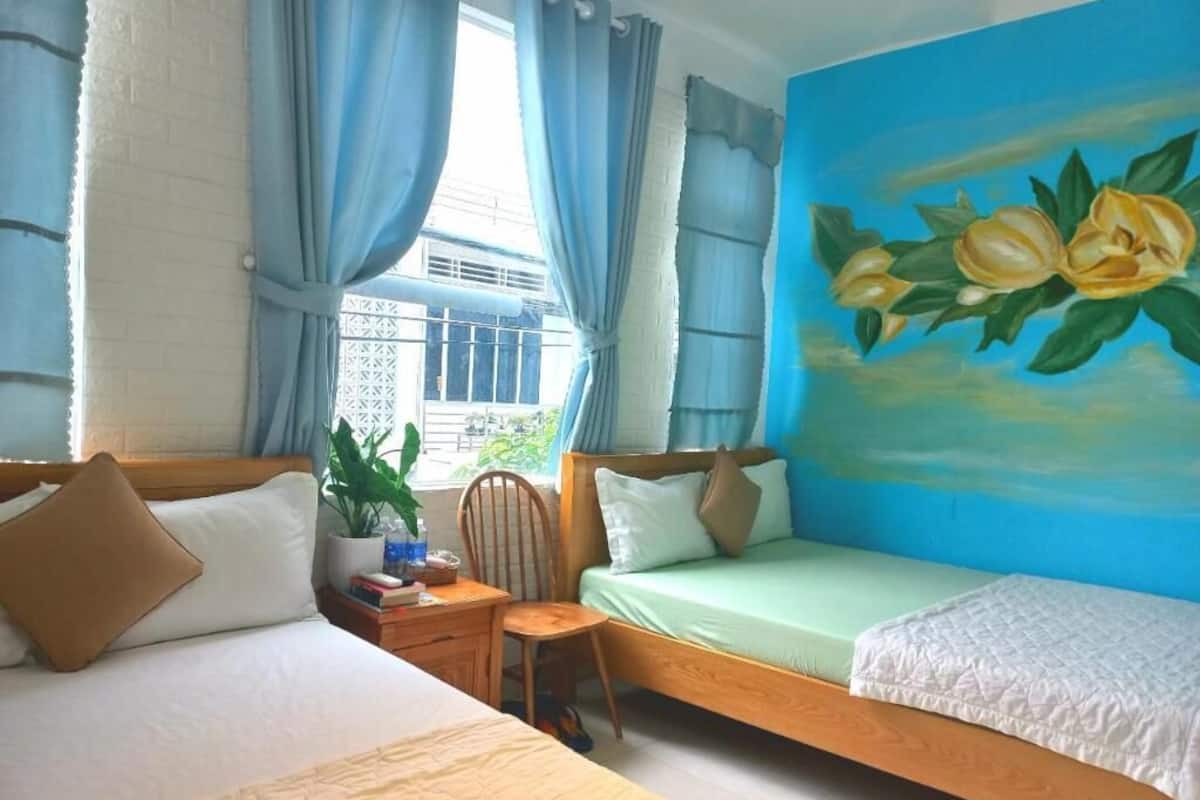 A bedroom with two beds, a window with curtains, a painting of flowers, and a potted plant.