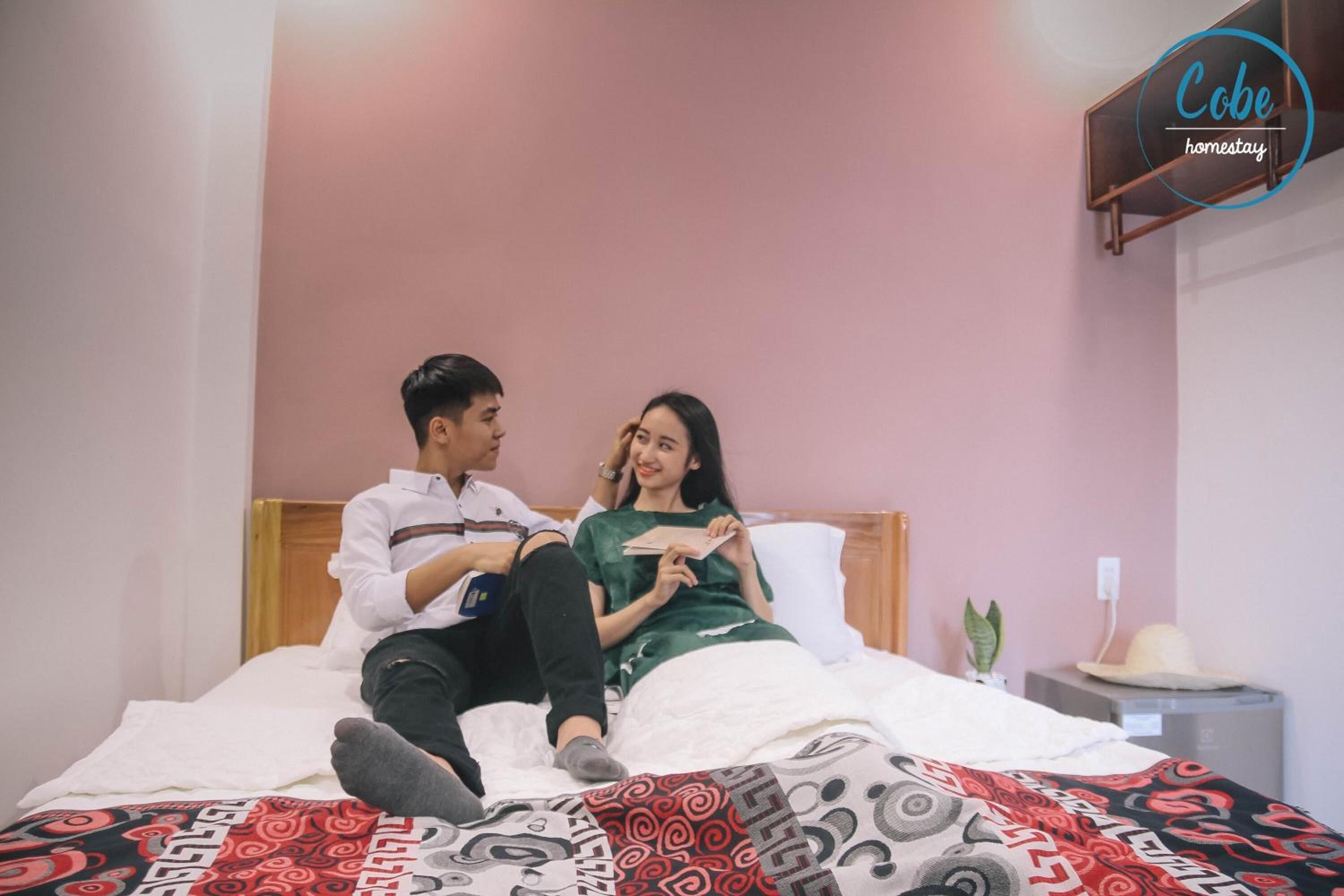 A couple sitting on a bed in a room with a pink wall, a bedside table with a hat, and a small plant.