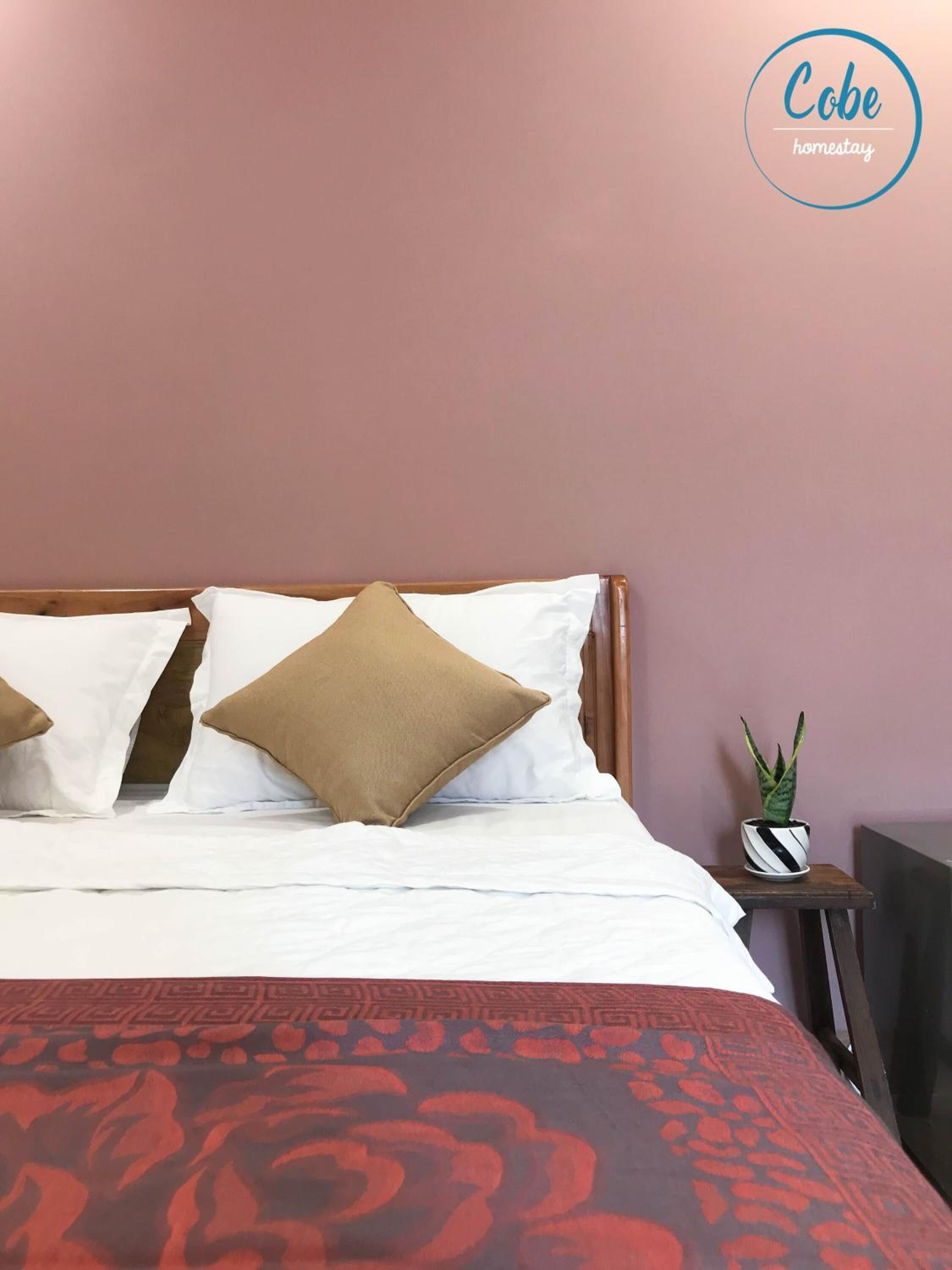 Uma cama bem arrumada com almofadas brancas e castanhas, um cobre-leito estampado em vermelho, e uma pequena planta em vaso sobre uma mesa de cabeceira ao lado da cama.
