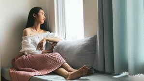 Una persona sentada en un banco junto a una ventana con cortinas, vestida con una blusa blanca de un hombro y una falda rosa.