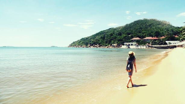 Una playa con una persona caminando por la orilla, una montaña al fondo y un edificio de techo rojo.