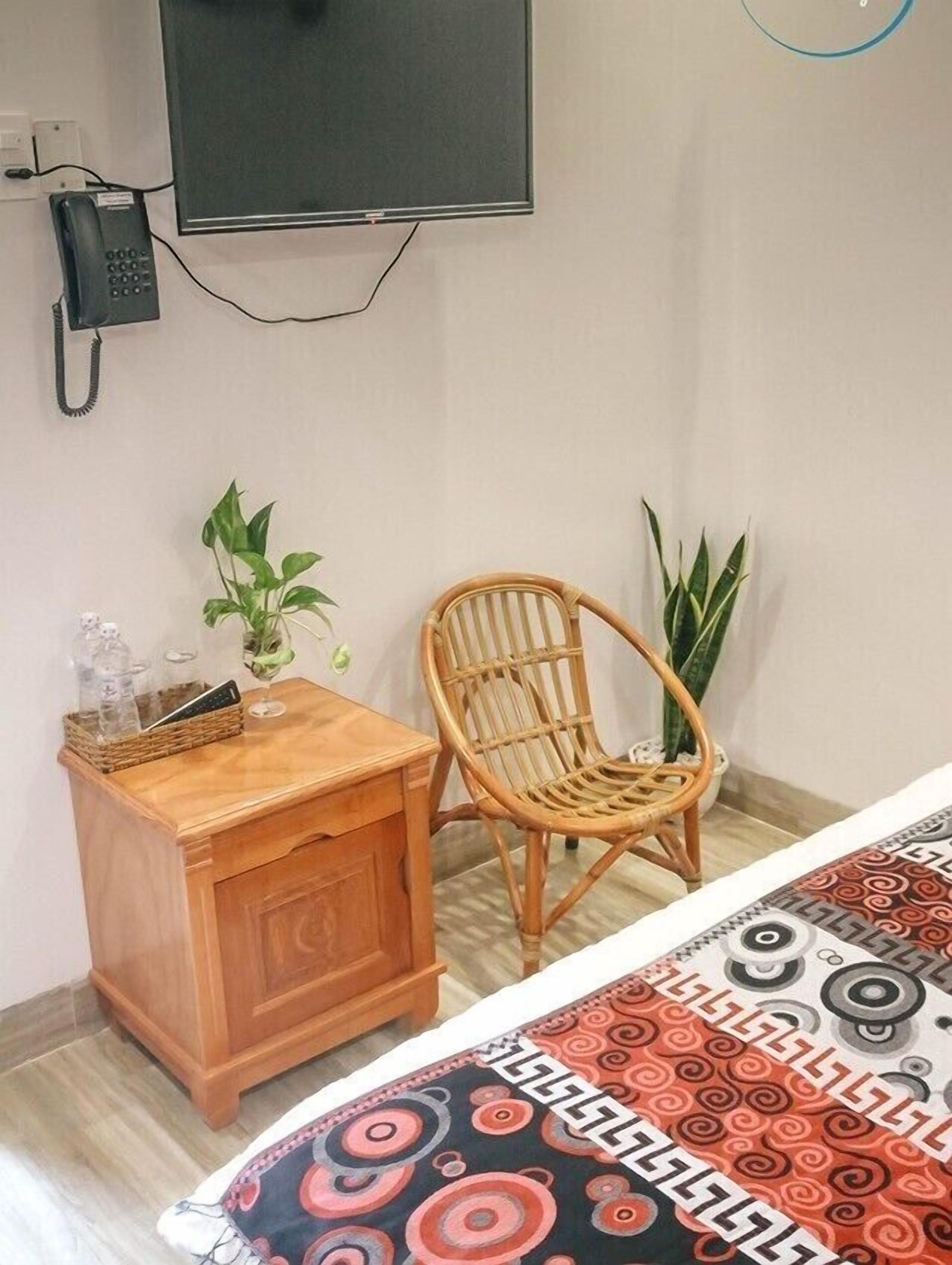 A hotel room with a bed, a wooden nightstand, a wicker chair, a potted plant, and a wall-mounted TV.