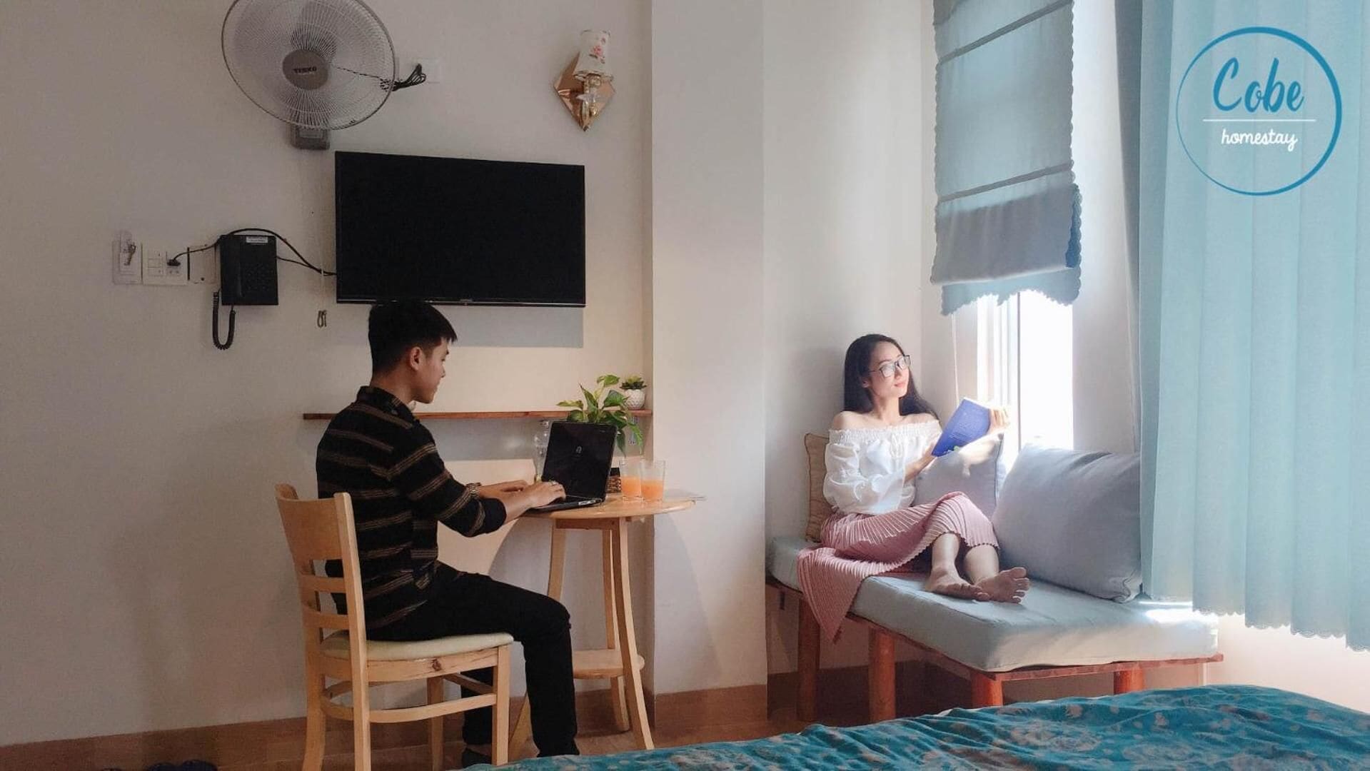 A room with a bed, a desk with a laptop, a chair, a TV on the wall, and a window with curtains.