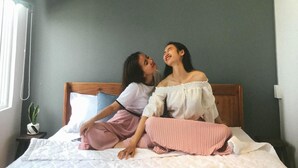 Un dormitorio con cama, ventana con cortinas, planta en la mesita de noche y lámpara en el techo.