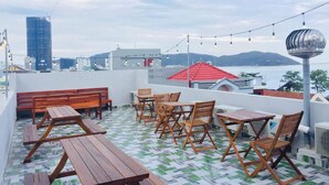 Terraza en la azotea con mobiliario de madera y luces colgantes, con vistas a un paisaje urbano y al agua.