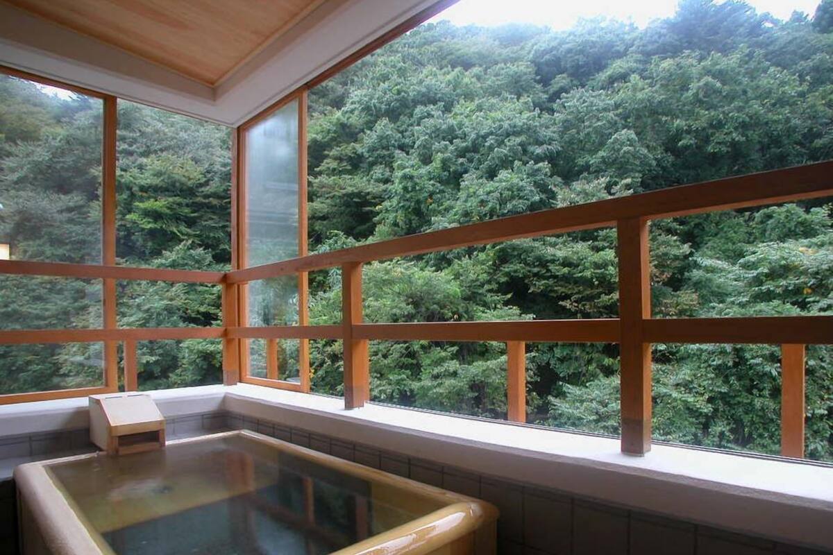 A room with a wooden bathtub and large windows overlooking a forest.