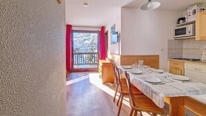 A spacious room with a wooden dining table set for four, a kitchen area with modern appliances, and a balcony with a scenic mountain view. - 100m from ski lifts, 3rd floor, balcony, tv, ski locker, 30m², Les Menuires (Les Menuires)