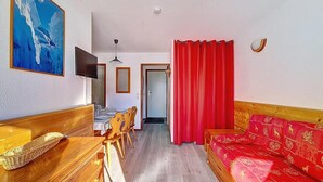 A room with a wooden bench, a red sofa, a dining table with chairs, a television, and a red curtain. - 100m from ski lifts, 3rd floor, balcony, tv, ski locker, 30m², Les Menuires (Les Menuires)