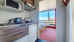 A compact kitchen with a microwave, coffee maker, and a small refrigerator. A window leads to a balcony with a bed and a view of buildings. - 100m from ski slopes, 7th floor, balcony, tv, ski locker, 30m², Les Menuires (Les Menuires)