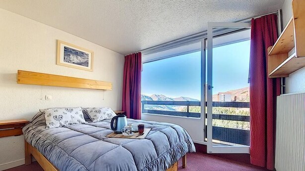 A bedroom with a bed, a window with a view of mountains, a wooden shelf, and a framed picture on the wall. - 100m from ski slopes, 7th floor, balcony, tv, ski locker, 30m², Les Menuires (Les Menuires)