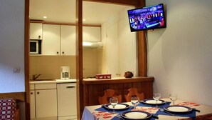Un área de comedor con una mesa dispuesta para cuatro, un televisor montado en la pared y una cocina al fondo.