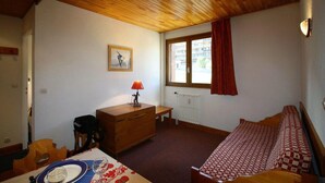 Un dormitorio con techo de madera, una cama con colcha a cuadros, un aparador de madera, una mesa con un vaso y una ventana con vistas a edificios.