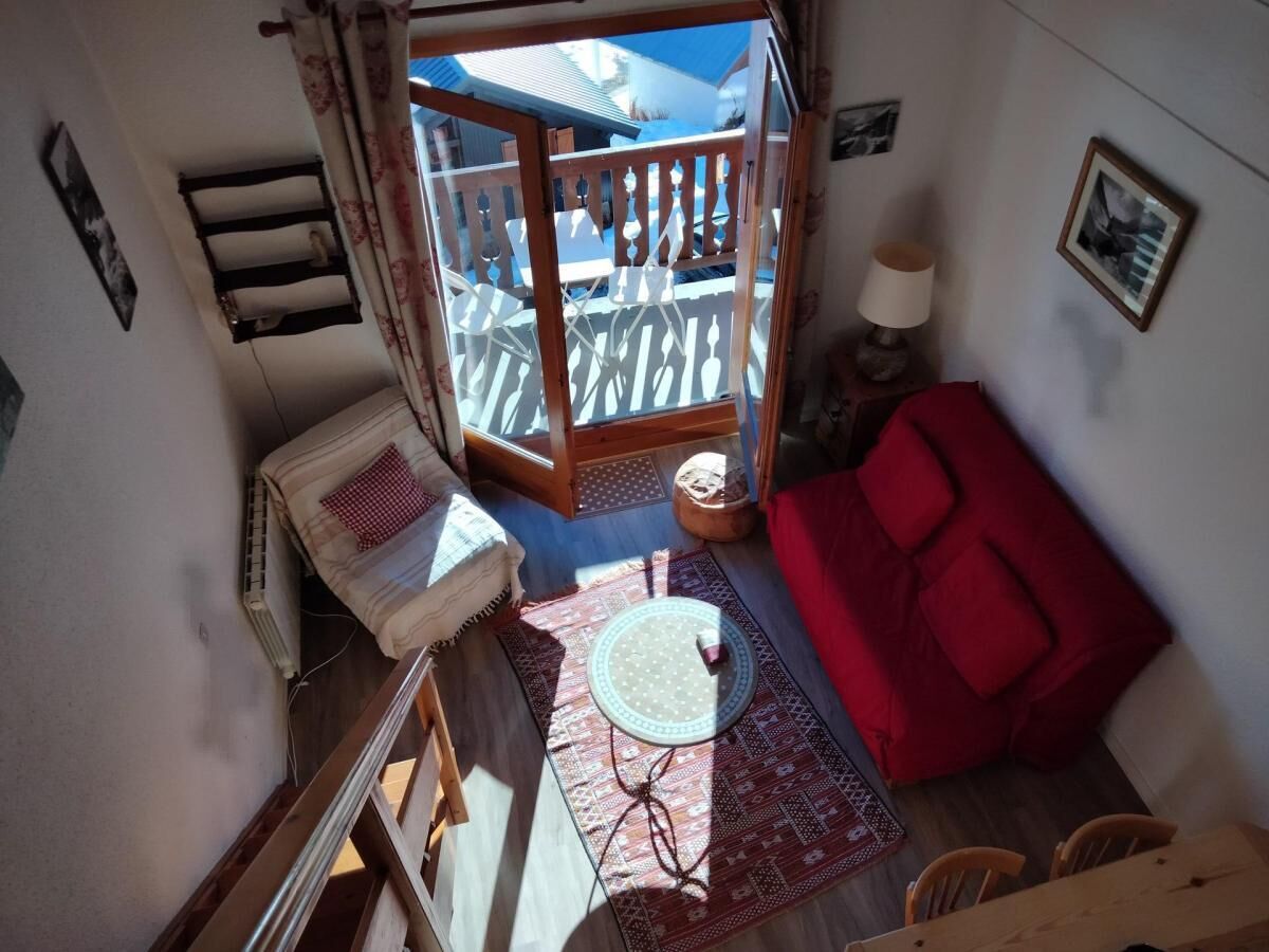 Een kleine kamer met een rode bank, een ronde tafel met een vloermat, een houten stoel, een tafeltje met een lamp, en uitzicht op een balkon met een leuning.