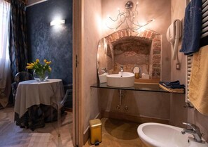 A bathroom with a glass-enclosed sink, a visible brick wall, a towel rack, and a small table with a vase of flowers. - Residenza San Faustino (Verona)