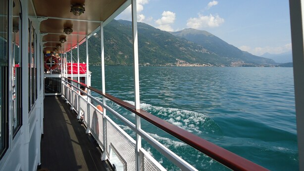 Ein Bootsdeck mit Metallgeländer, einem überdachten Gang und Blick auf einen See mit Bergen im Hintergrund.