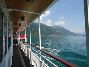 Ein Bootsdeck mit Metallgeländer, einem überdachten Gang und Blick auf einen See mit Bergen im Hintergrund.