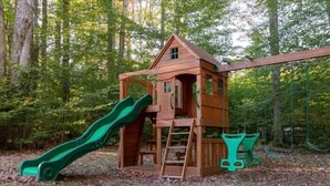 Ein hölzernes Spielhäuschen mit Rutsche und Kletterelementen, umgeben von einem Wald.