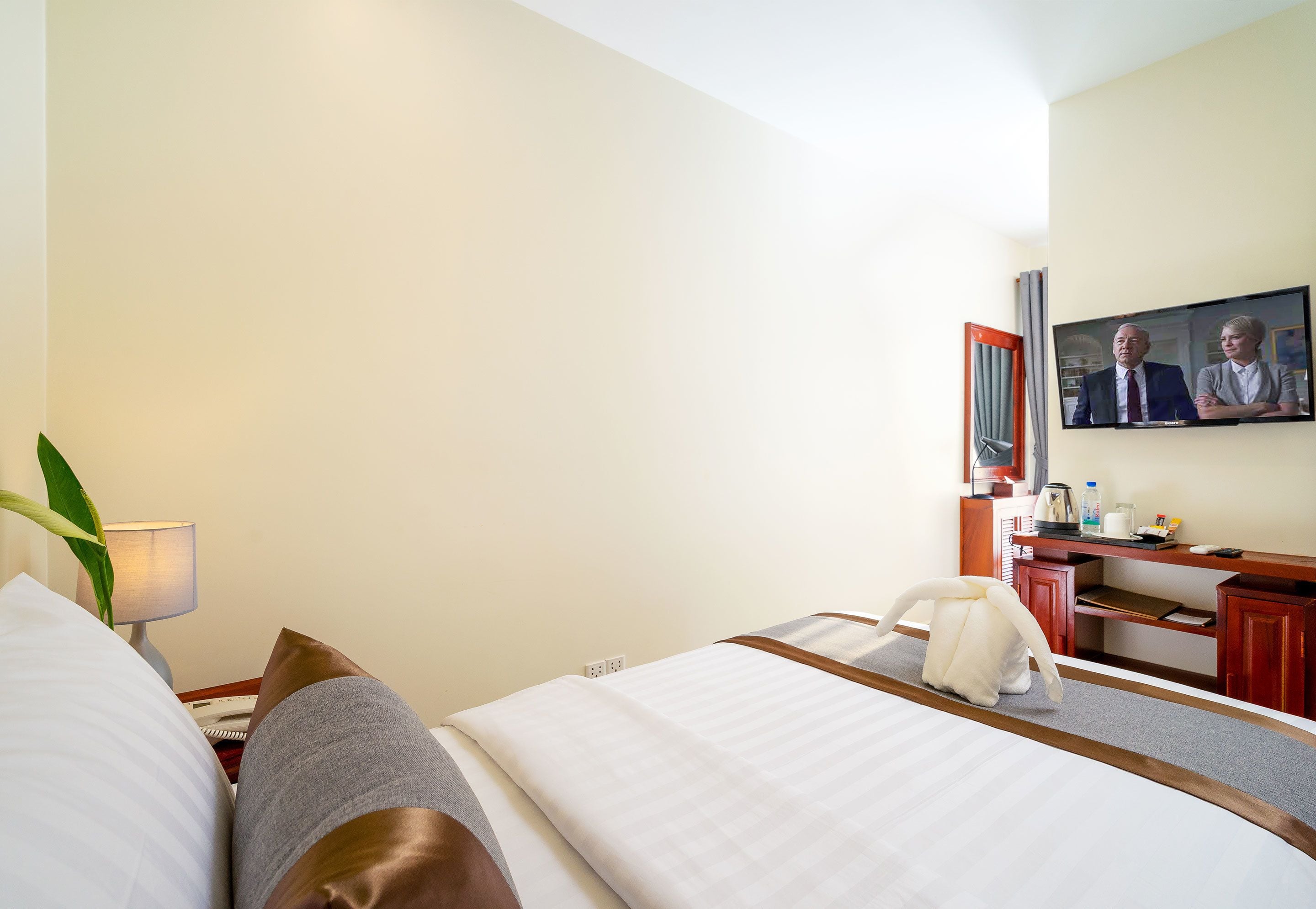 Een hotelkamer met een bed, nachtkastje, lamp en een televisie waarop twee personen te zien zijn.