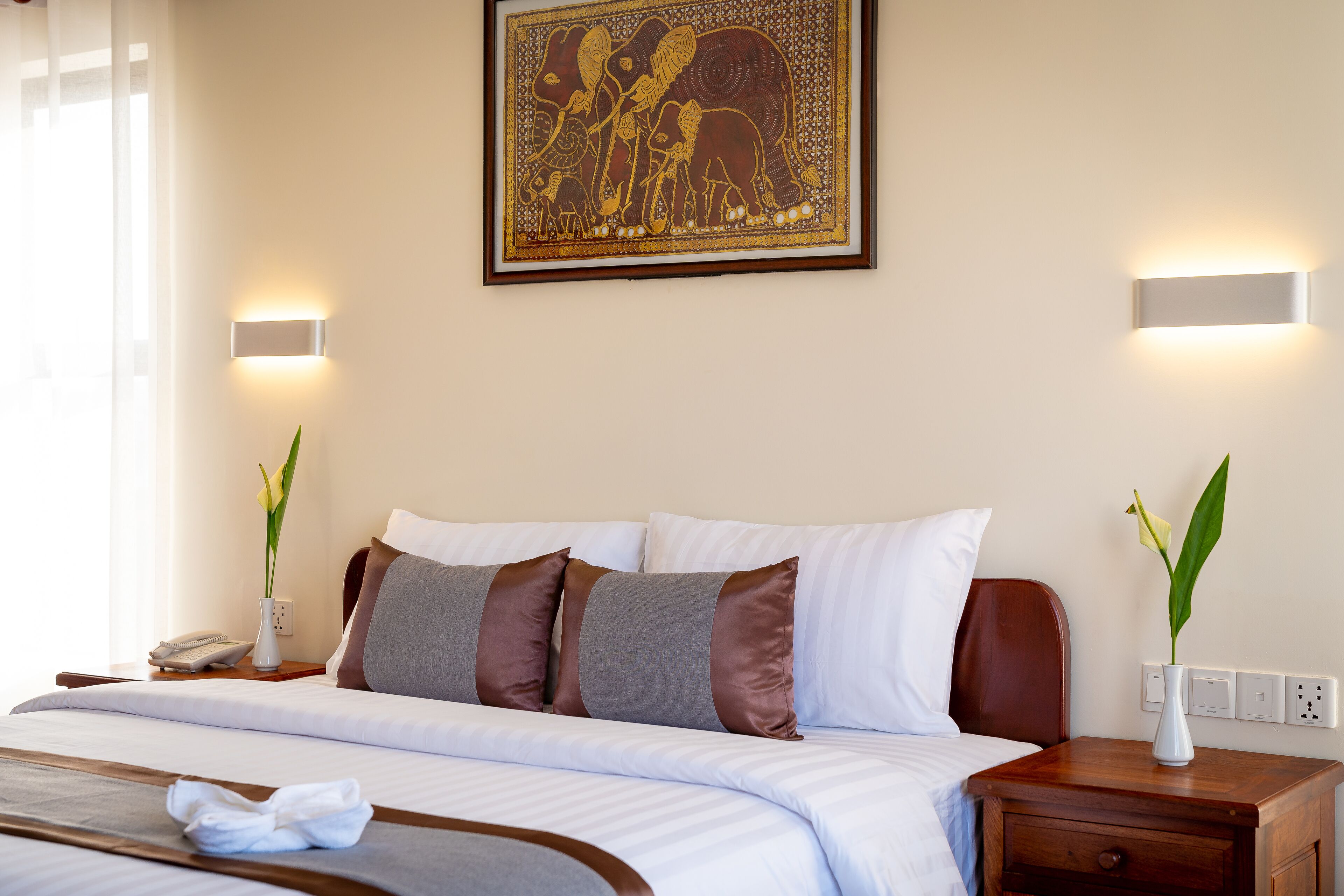 Habitación de hotel con una cama bien hecha, una mesita de noche con lámpara, un jarrón con flores y una obra de arte colgada en la pared.