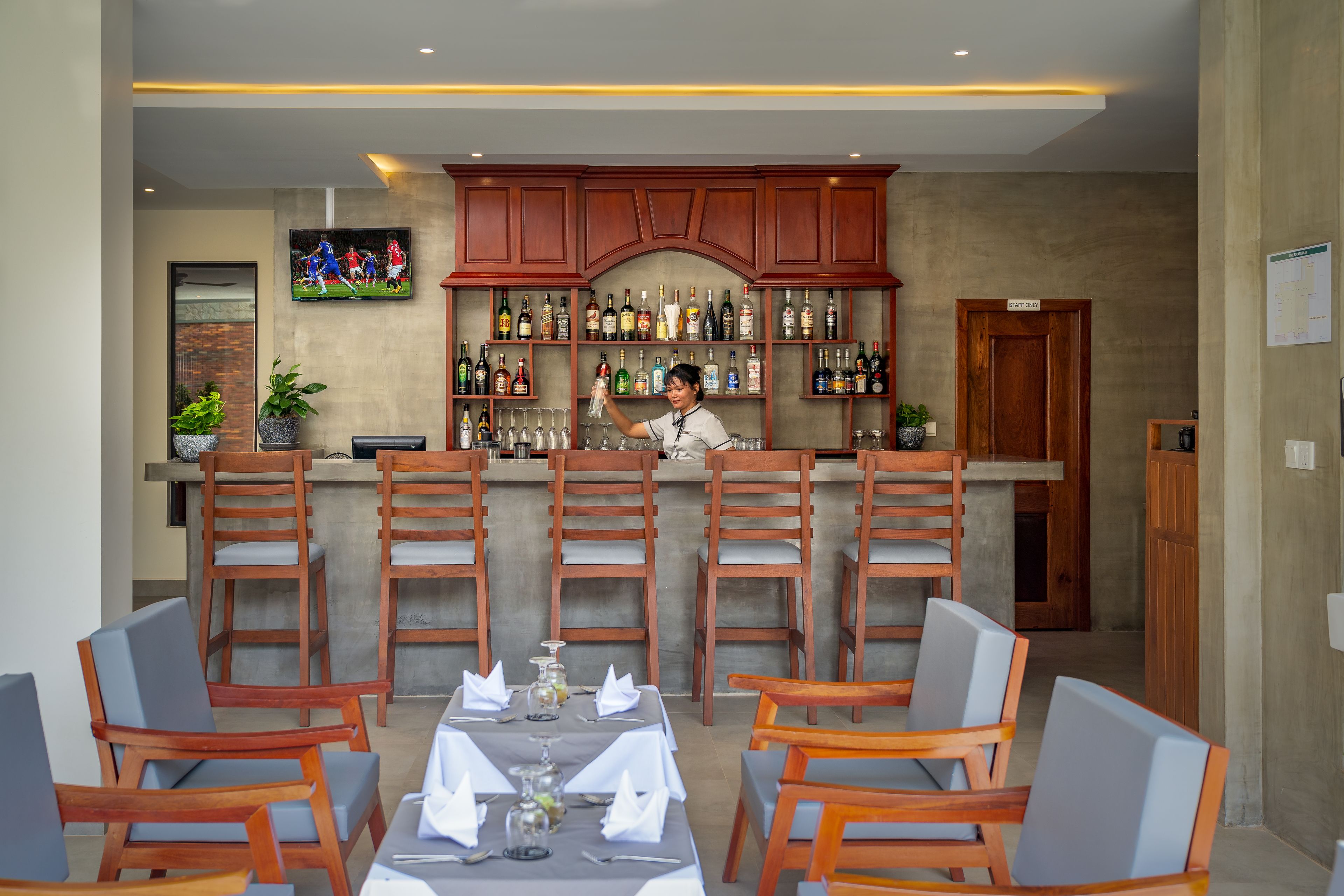A restaurant with wooden chairs and tables, a bar area with shelves of bottles, and a television mounted on the wall.