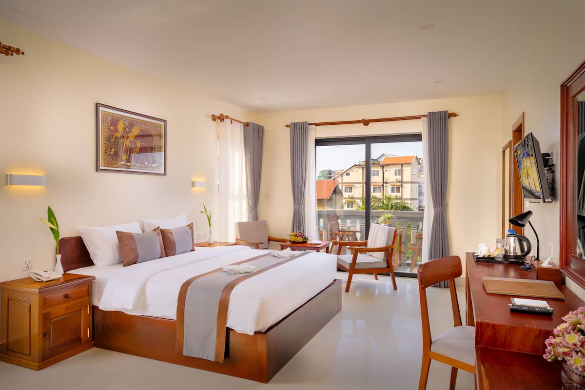 A hotel room with a large bed, a desk with a chair, a balcony with a view of buildings, and a painting on the wall.