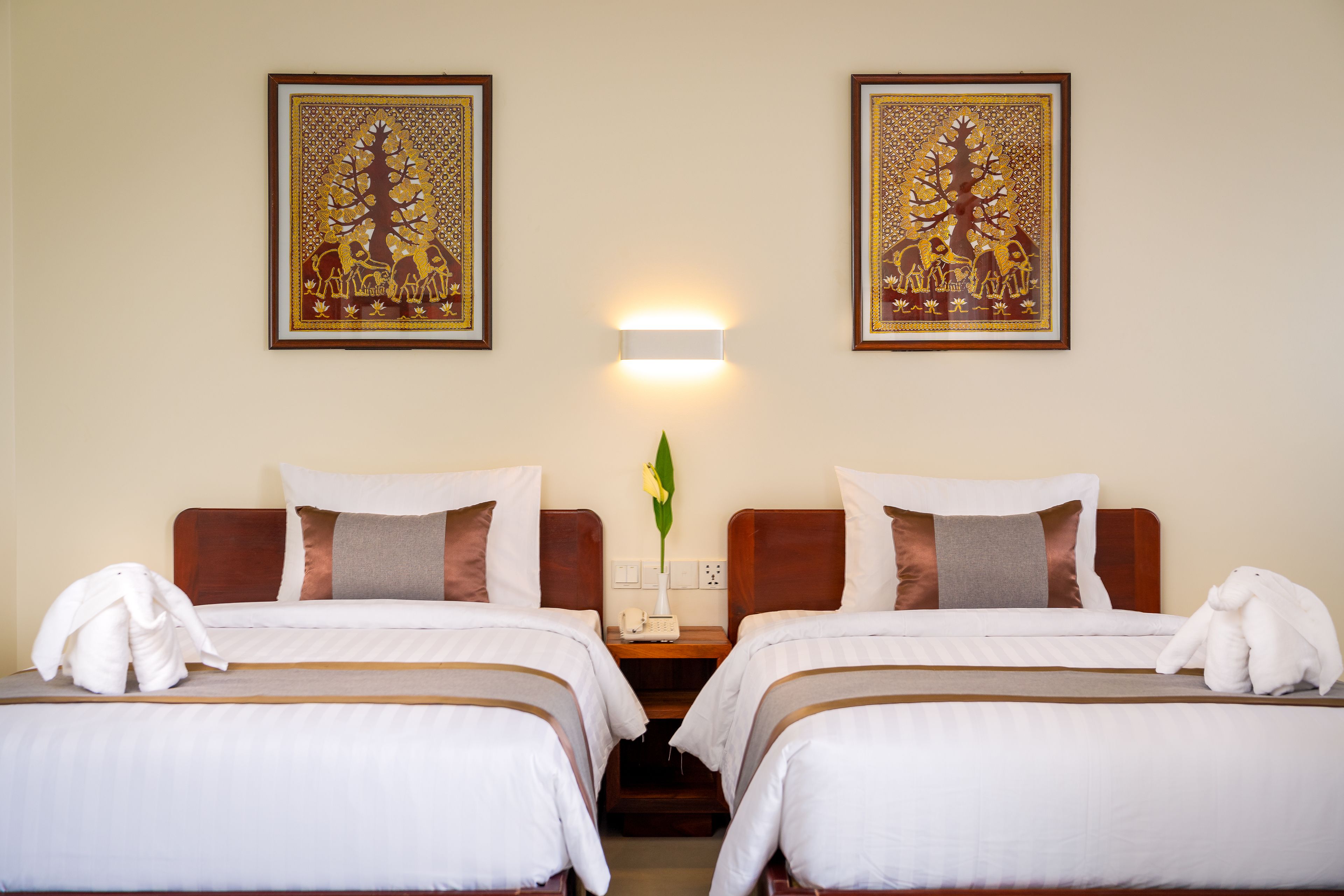 A hotel room with two beds, a nightstand with a lamp, and two framed artworks on the wall.